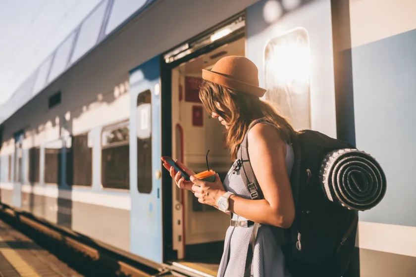 Young woman with a backpack and hat checks her phone on a platform beside an open train door in the sunlight